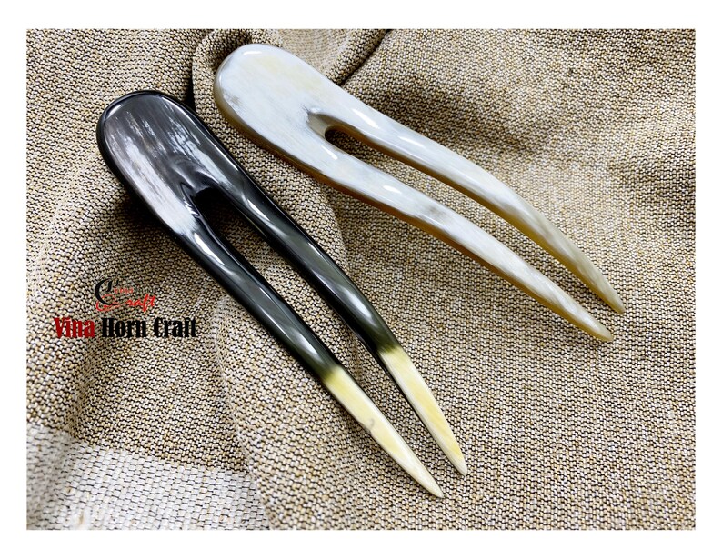 Puede incluir: Dos horquillas para el cabello hechas de cuerno. Una es gris oscuro y la otra blanquecina. Ambas tienen un acabado liso y pulido y un dise&ntilde;o de dos puntas. Las puntas de las horquillas son de un color m&aacute;s claro, creando un efecto degradado. Las horquillas se exhiben sobre una tela beige texturizada.