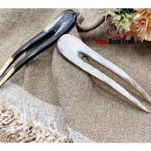 Puede incluir: Dos horquillas para el cabello hechas de cuerno. Una es gris oscuro con puntas amarillas, la otra es blanquecina. Ambas est&aacute;n sobre una tela beige texturizada. Accesorios para el cabello.