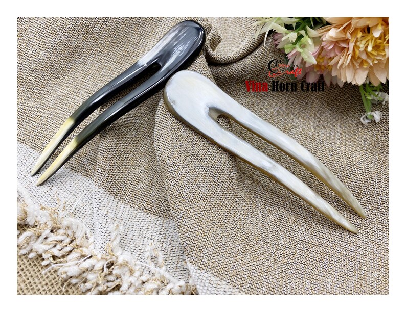 Puede incluir: Dos horquillas para el cabello hechas de cuerno. Una es gris oscuro con puntas amarillas, la otra es blanquecina. Ambas est&aacute;n sobre una tela beige texturizada. Accesorios para el cabello.