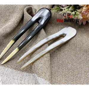 Puede incluir: Dos horquillas para el cabello de cuerno. Una es gris oscuro con punta amarilla, la otra blanquecina. Ambas tienen un dise&ntilde;o en forma de U. Las horquillas se muestran sobre una tela texturizada, de color beige. El texto "Vina Horn Craft" es visible.