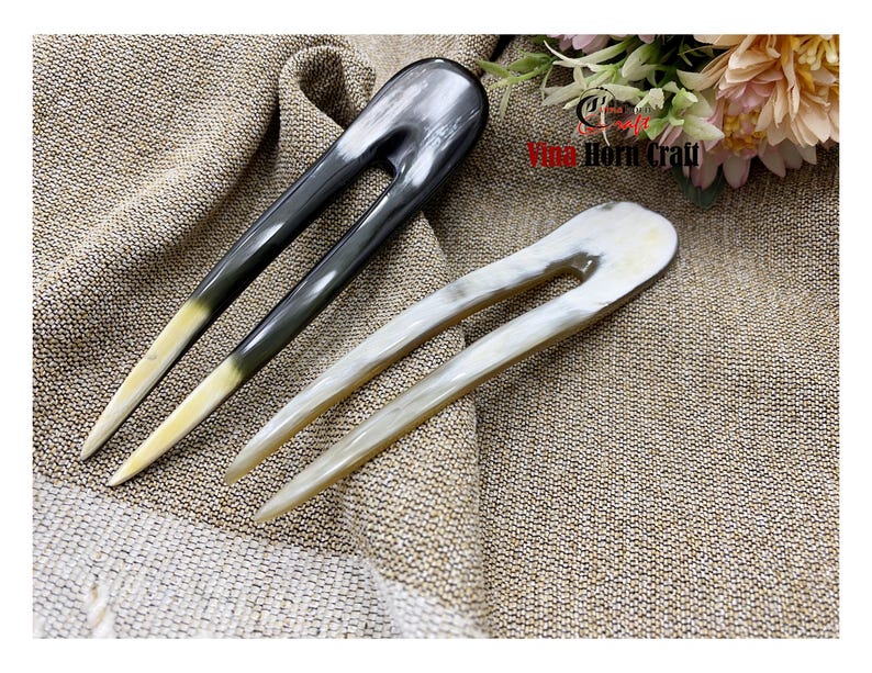 Puede incluir: Dos horquillas para el cabello de cuerno. Una es gris oscuro con punta amarilla, la otra blanquecina. Ambas tienen un dise&ntilde;o en forma de U. Las horquillas se muestran sobre una tela texturizada, de color beige. El texto "Vina Horn Craft" es visible.