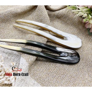 Puede incluir: Dos horquillas para el cabello de cuerno. Una es blanca, la otra negra con punta blanca. Las horquillas est&aacute;n sobre una superficie beige texturizada. El texto "Vina Horn Craft" es visible en la esquina inferior izquierda.