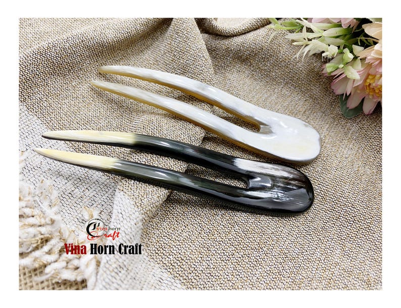 Puede incluir: Dos horquillas para el cabello de cuerno. Una es blanca, la otra negra con punta blanca. Las horquillas est&aacute;n sobre una superficie beige texturizada. El texto "Vina Horn Craft" es visible en la esquina inferior izquierda.