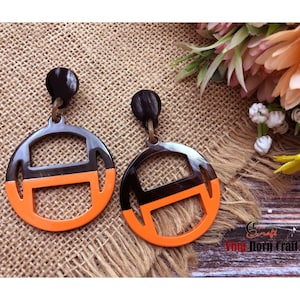 Może przedstawiać: Para czarno-pomarańczowych okrągłych kolczyków z wyciętym wzorem. Kolczyki są wykonane z rogu i mają czarno-pomarańczową kolorystykę. Kolczyki wiszą na brązowym tle z juty.