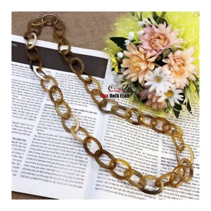 May include: A brown and white horn chain necklace. The chain is made of oval links. The necklace is lying on a book with text in a foreign language. The book is on a yellow surface with a bouquet of flowers.