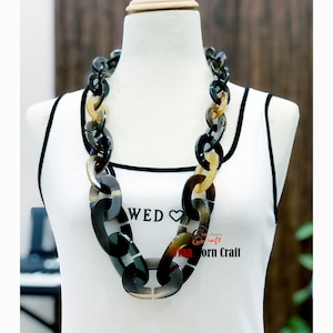 May include: A large, chunky necklace made of brown and black horn. The necklace is on a white mannequin and has a 'WED' and heart design on the front of the mannequin's shirt. The text 'Vina Horn Craft' is also visible on the shirt.