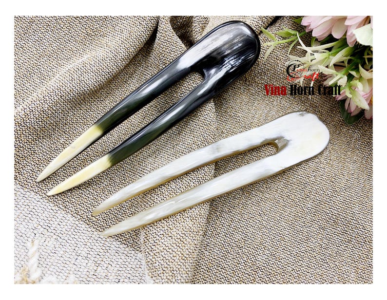 Puede incluir: Dos horquillas para el cabello de cuerno. Una es negra y la otra blanca. Ambas tienen forma de U con dos p&uacute;as largas y puntiagudas. Las puntas de las horquillas son de color crema claro. Las horquillas se muestran sobre una superficie texturizada y marr&oacute;n.