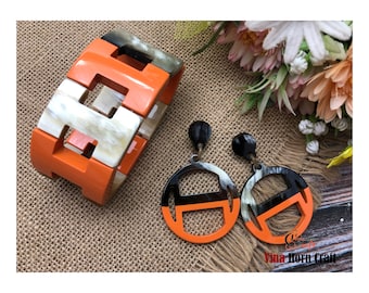 May include: An orange and cream-colored bracelet and matching earrings set. The bracelet is made of rectangular blocks with cut-out designs. The earrings are circular with a similar cut-out design, featuring black and orange accents. The jewelry set is displayed on a textured surface.