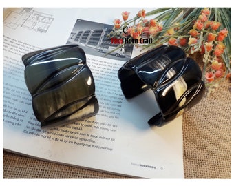 May include: Two black and white horn bracelets with a carved design. The bracelets are on a brown surface with a book and flowers in the background.