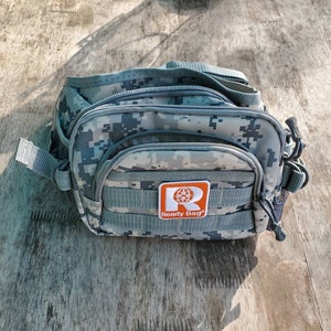 Puede incluir: Una riñonera de camuflaje gris con una cremallera negra y un parche naranja que dice "Ready Bag" con un icono de globo terráqueo.
