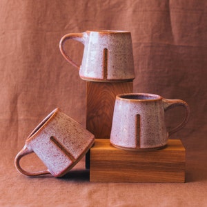 May include: Three ceramic mugs with a speckled pink and brown glaze, each with a brown vertical stripe and handle. The mugs are displayed on wooden blocks against a brown backdrop. The mugs are a unique addition to any kitchen.