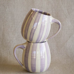 A stack of two handmade ceramic mugs with light purple vertical stripes painted on the white clay