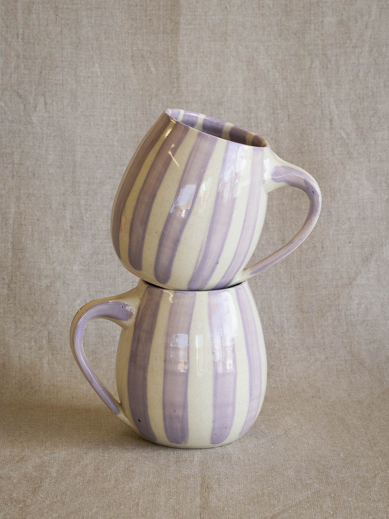 A stack of two handmade ceramic mugs with light purple vertical stripes painted on the white clay