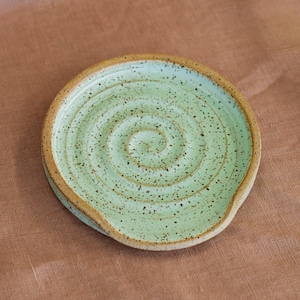 May include: A handmade ceramic spoon rest with a light green glaze and a spiral design. The spoon rest has a speckled appearance and a brown rim. The design is functional and decorative, perfect for the kitchen.