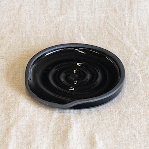 May include: A black ceramic spoon rest with a glossy finish. The spoon rest has a circular design with concentric rings and a small lip on one side. The spoon rest is on a neutral-colored surface.