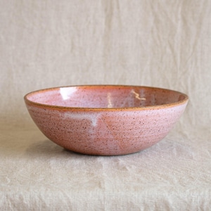 May include: A speckled, pink ceramic bowl with a wide, shallow design. The rim has a darker, reddish-brown color. The bowl is likely handmade and suitable for serving food or as a decorative piece.