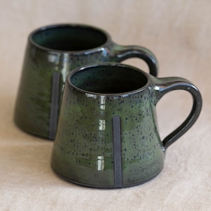 May include: Two handmade ceramic mugs with a dark green glaze and black accents. Each mug has a unique shape with a handle and a vertical black stripe. The mugs are speckled and have a glossy finish.