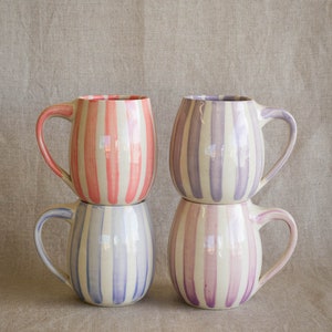 Four handmade ceramic mugs stacks together, each with vertical hand painted stripes in pink, lavender, periwinkle, and purple