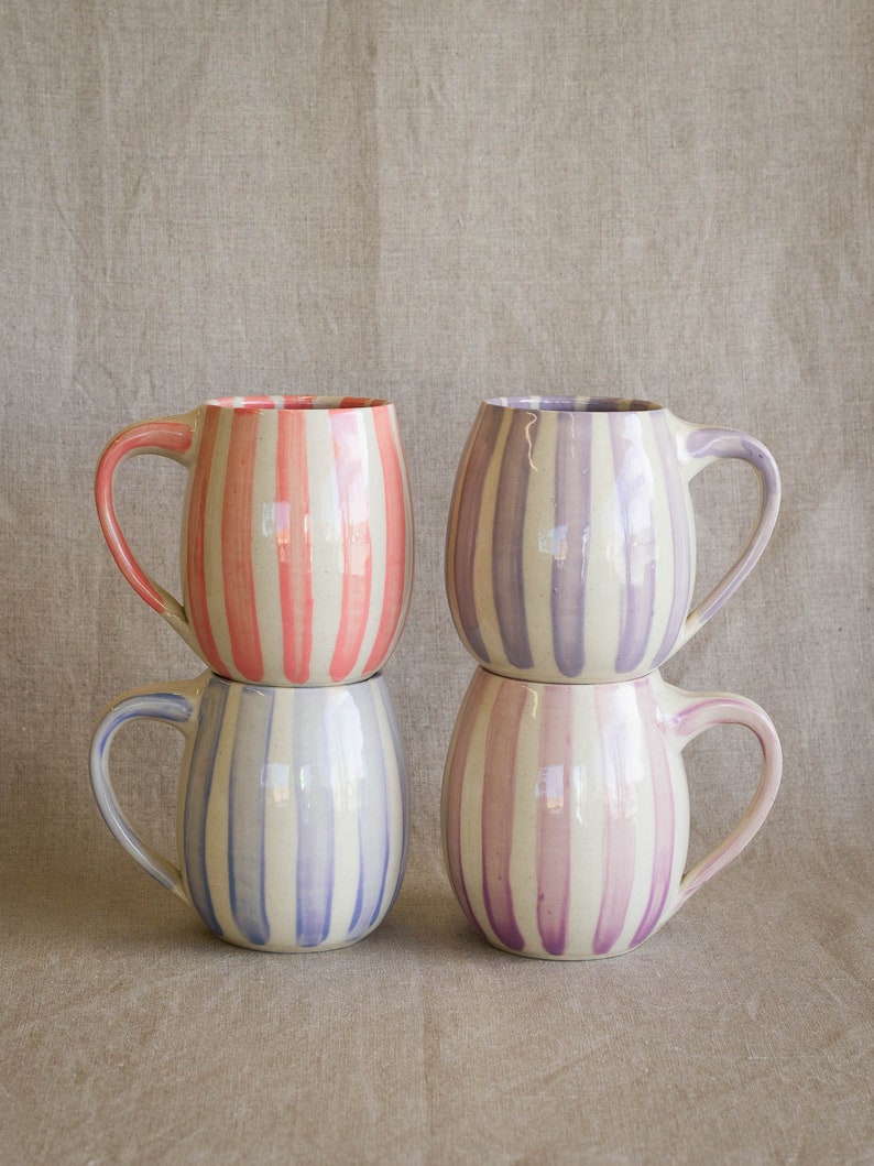 Four handmade ceramic mugs stacks together, each with vertical hand painted stripes in pink, lavender, periwinkle, and purple