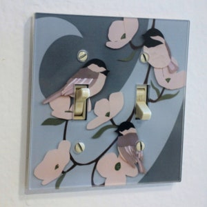 May include: A light switch cover with a blue background and a design of pink flowers and brown birds. The switch cover has two toggle switches.