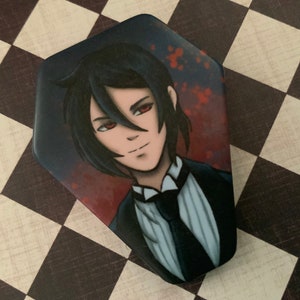 May include: A coffin-shaped decorative item featuring an anime-style illustration of a character with black hair and red eyes. The character is dressed in a black suit with a white shirt and black tie. The background has a red and black color scheme.