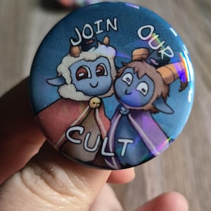 May include: A round button with a blue background and the words "JOIN OUR CULT". The button features two cartoon goat-like characters with red and purple capes. The button has a holographic effect.