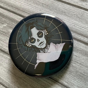 May include: A black and gold button featuring a stylized illustration of a person with a skull face and dark hair. The person is wearing a white garment with a black background.