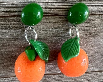 May include: A pair of orange and green fruit-themed earrings. Each earring features a bright orange fruit with green leaves and a green top, connected by a silver-coloured loop. The earrings are set against a wooden background.