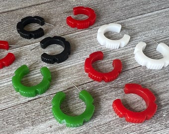 May include: A selection of hoop earrings in red, black, white, and green. The earrings feature a textured, scalloped design. The earrings are displayed on a light grey wooden surface.
