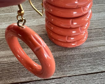 May include: Coral-coloured ceramic earrings with gold-coloured hooks. The earrings are a rounded, oval shape with a textured design. A stack of matching coral-coloured napkin rings is also visible.