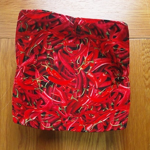 May include: A square bowl cosy made of red fabric, featuring a repeating pattern of red chilli peppers against a black backdrop. The cosy is open at the top and rests on a wooden surface.