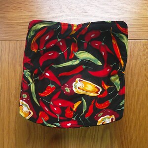 May include: A black fabric item featuring a colourful pattern of red, yellow, and green chilli peppers. The peppers are spread across the surface, creating a striking design. The item appears to be a kitchen accessory, perhaps a pot holder.