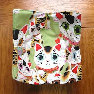 May include: A square, green fabric item with a repeating pattern of Japanese Maneki Neko cats. The cats are white with red ears, black markings, and colourful details. The background is a light green.