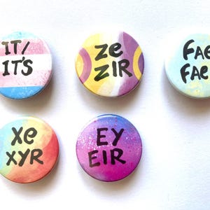 Op de afbeelding: Zes ronde knoppen met verschillende pronomensets erop gedrukt. De knoppen zijn kleurrijk en hebben een regenboog-, blauw-, geel-, paars- en groenschema. De pronomen zijn "it/its", "ze/zir", "fae/faer", "xe/xyr", "ey/eir" en "they/them".