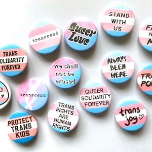 Könnte beinhalten: Eine Sammlung von 14 runden, bunten Buttons mit verschiedenen Botschaften zur Förderung von Transgender-Rechten und Solidarität. Die Buttons zeigen Sätze wie "Trans Power", "Trans Rights Are Human Rights", "Queer Solidarity Forever" und "Always Been Here".
