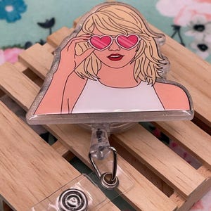 Könnte beinhalten: Ein durchsichtiger Plastik-Badgehalter mit Glitzer-Hintergrund, der eine Comic-Illustration einer Frau mit herzförmigen Sonnenbrillen zeigt. Der Badgehalter ist an einer einziehbaren Badge-Spule befestigt.