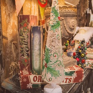 May include: A white, frosted, cone-shaped Christmas tree with green holly and red berries. The tree is decorated with white glitter and is sitting on a table with a variety of vintage Christmas decorations, including books with the words "Happy Holiday" and "Merry Christmas" on the covers.