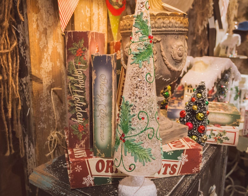 May include: A white, frosted, cone-shaped Christmas tree with green holly and red berries. The tree is decorated with white glitter and is sitting on a table with a variety of vintage Christmas decorations, including books with the words "Happy Holiday" and "Merry Christmas" on the covers.