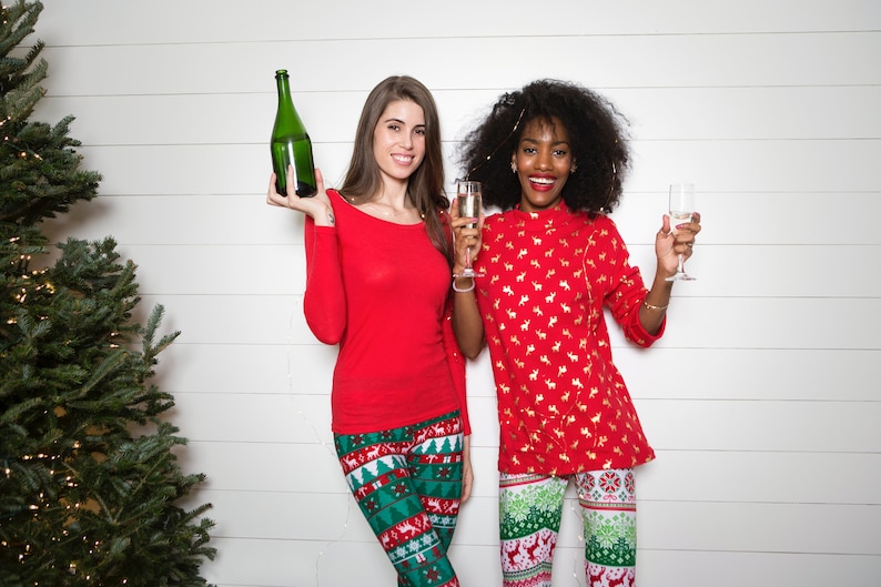 May include: Two people wearing red and green Christmas pajamas are holding a bottle of champagne and two glasses. They are standing in front of a white wood wall with a Christmas tree to the left.