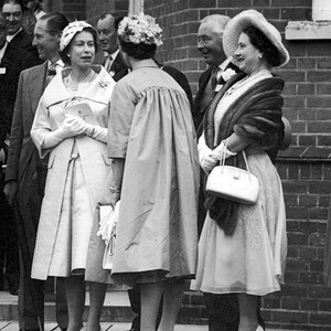 May include: Black and white photograph of a group of people, including several women in dresses and hats. One woman wears a light-colored coat and holds a small bag. The group is standing outside near a brick building.