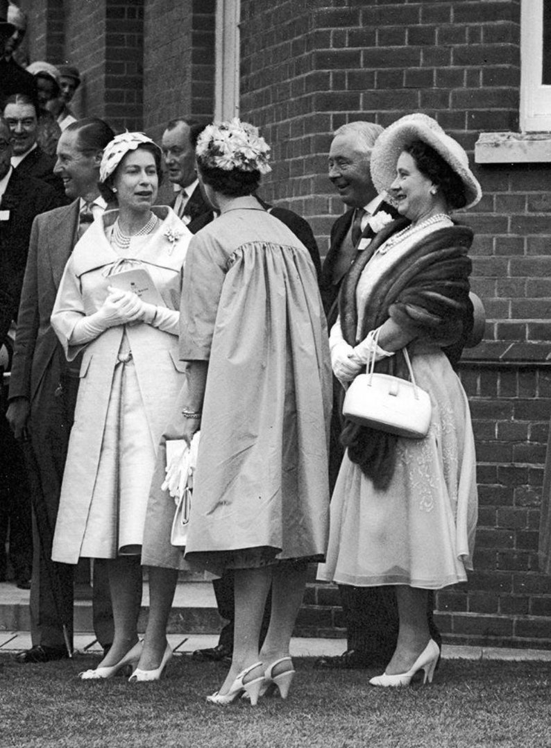 May include: Black and white photograph of a group of people, including several women in dresses and hats. One woman wears a light-colored coat and holds a small bag. The group is standing outside near a brick building.
