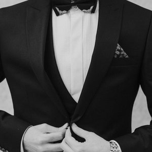 May include: A man wearing a black tuxedo with a white shirt and a black bow tie. He is wearing a patterned pocket square and a silver watch.