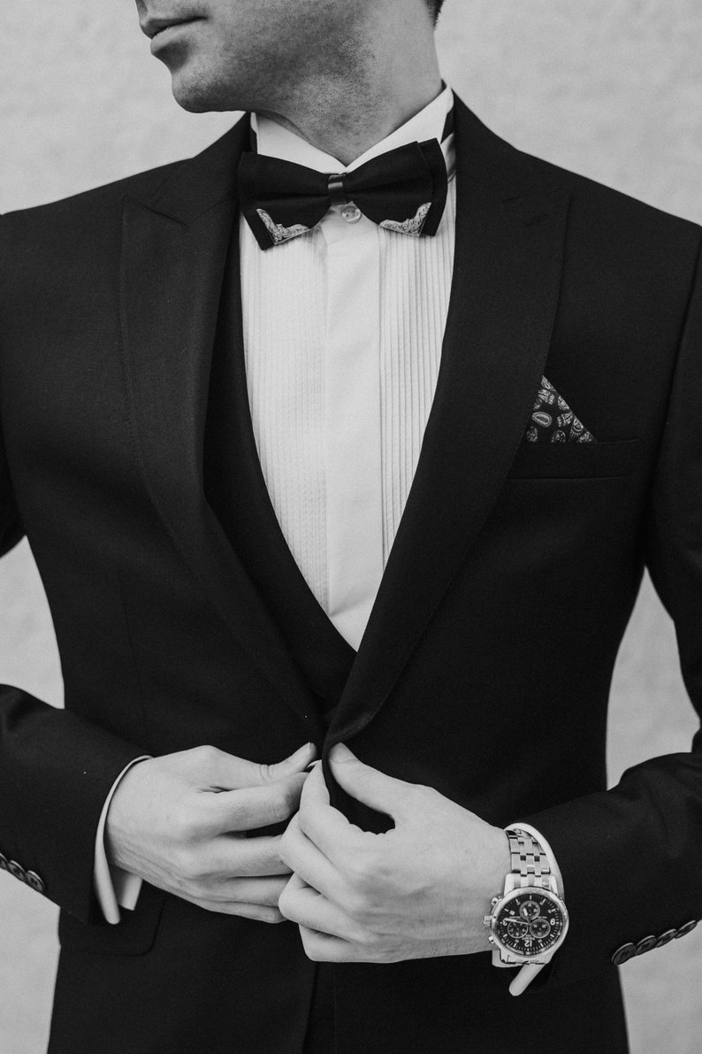 May include: A man wearing a black tuxedo with a white shirt and a black bow tie. He is wearing a patterned pocket square and a silver watch.