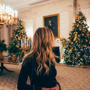 May include: A woman in a black dress stands in a room decorated for the holidays.  Two Christmas trees are visible in the background, one on each side of the woman.  The trees are decorated with lights and ornaments.  The room is lit with warm, inviting light.