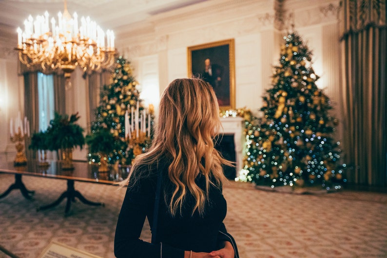 May include: A woman in a black dress stands in a room decorated for the holidays.  Two Christmas trees are visible in the background, one on each side of the woman.  The trees are decorated with lights and ornaments.  The room is lit with warm, inviting light.