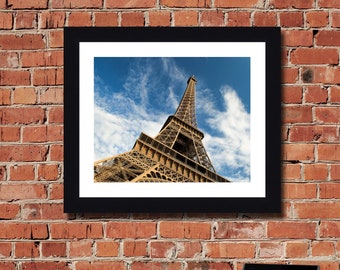 May include: A framed photograph of the Eiffel Tower in Paris, France. The tower is made of metal and is a light brown color. The sky is blue with white clouds. The photo is framed in black.