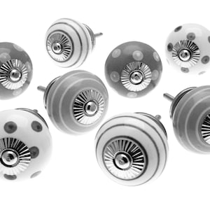 Op de afbeelding: Een verzameling van acht decoratieve keramische kastknoppen. De knoppen hebben verschillende ontwerpen, waaronder stippen en strepen, in grijstinten en wit. Elke knop heeft een zilverkleurige metalen basis en steel.