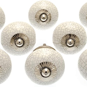 Könnte beinhalten: Acht weiße Keramik-Schrankknöpfe mit einem Craquelé-Glasurmuster. Jeder Knopf hat einen silberfarbenen Metallsockel und eine zentrale Schraube. Diese dekorativen Knöpfe eignen sich für Möbel und Heimdekorationsprojekte.