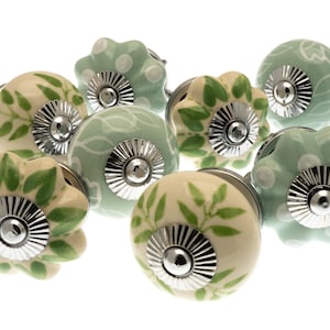 May include: A collection of ceramic cabinet knobs in various designs. The knobs feature floral and leaf patterns in shades of green and cream, with silver-toned metal bases. Some have a polka dot design.