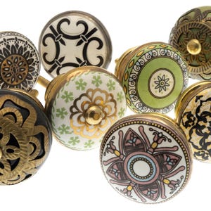 Puede incluir: Una colección de siete pomos redondos de cerámica para armarios con diversos patrones decorativos en negro, blanco, verde, dorado y rosa. Cada pomo tiene una base metálica y un diseño circular único, incluyendo motivos florales y geométricos.